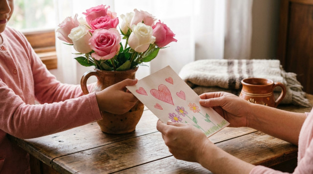 ramo de rosas y tarjeta hecha a mano sobre una mesa de madera, con tonos rosa y blanco, luz natural suave, detalle de manos entregando la tarjeta