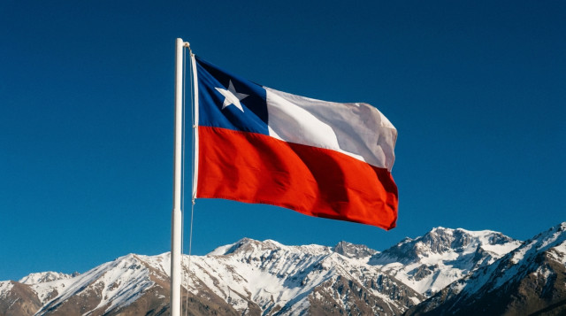 Bandera de Chile flameando en un mástil blanco bajo un cielo azul intenso con la Cordillera de los Andes de fondo