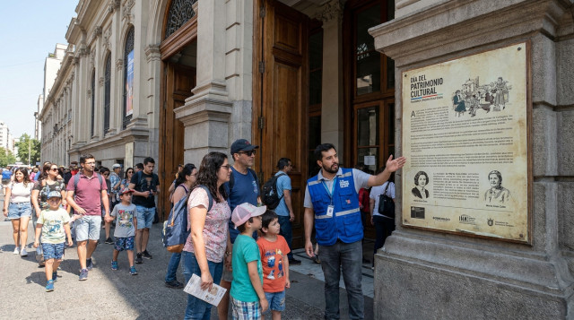 fachada de un edificio patrimonial chileno con gente entrando durante una jornada del Día del Patrimonio, detalle de cartel informativo y un guía explicando a una familia